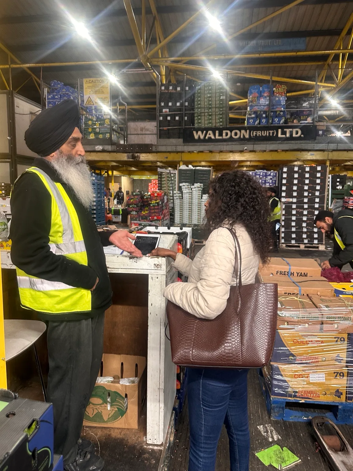 Doris Beth Atigo speaking with a trader beside fresh-produce boxes at Spitalfields Market.